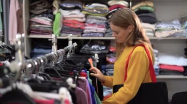 Portrait of young beautiful woman choosing clothes in store. stands near showcase with womens clothing sorting through different versions of pants on a hanger. The concept of shopping and consumerism
