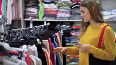 Portrait of a lady choosing clothes in a store. stands near the hangers holds a credit card in his hands and decides what to buy. The concept of shopping and consumerism.