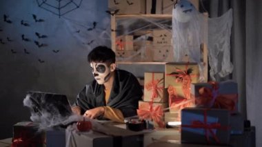 arab man in halloween make-up sits in home decorated office processes applications on website online. Boxes with orange ribbon and word sale concept of discounts and promotions for day of all the dead