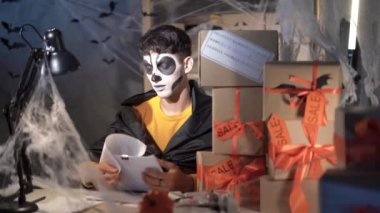 arab male business owner in halloween makeup sitting in home decorated office checking reports. Concept sale discounts and promotions for the day of all the dead