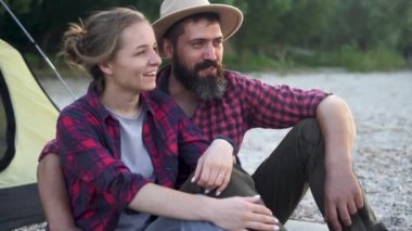 A young couple sits near a tent while relaxing in nature near a lake on the shore and looks at each other on a beautiful evening. Adventure and family travel concept.