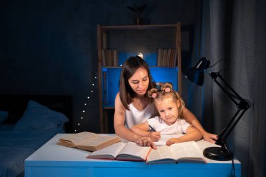 Mother and child daughter doing homework writing and reading at home in the evening. Copy space