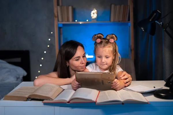 mom helps child daughter do homework at home using digital tablet. Child studying late at night. Children, family, education concept