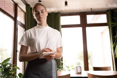attractive waitress in an apron standing in a cozy coffee shop, a cafe worker with a notebook in her hands ready to receive customers, ordering guests in a restaurant, copy space