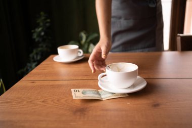 Woman hand taking cup wsth US dollar banknotes on cafe's table, two cups of tea on background. Concepts for payment, service charge, bill checking, money tips.