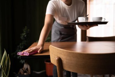 young waitress in apron bending over wooden table using duster to clean it for new guests of restaurant. Copy space
