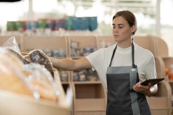 Confident supermarket female clerk working with a digital tablet. Young supervisor with tablet pc in the mall, background of bread shelves, technology and work concept.