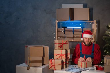 bearded man wearing a Santa Claus hat works in a warehouse of boxes with gifts and orders from an online store for Christmas. Small Business Owner Sells Happy New Year. Delivery holiday.