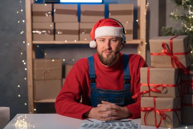 Smiling Young bearded Santa Claus wearing hat with headphones looking at camera sitting in workshop warehouse. Xmas delivery concept. Portrait. SME, e-commerce, telemarketing