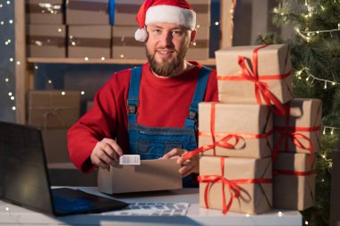 Caucasian business owner working with product packaging and laptop wearing a Sanat Claus hat and red sweater. Glue barcode on the box and prepares the purchase for shipment. Christmas sale