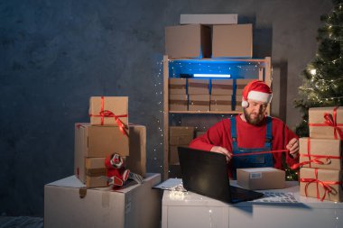 salesman in a Santa Claus hat takes an order by laptop while sitting in an office with boxes of gifts and surprises for Christmas for office workers. Shopping and delivery. Small business concept