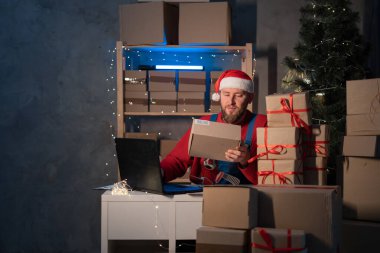 Santa Claus wearing hat holding gift box using laptop computer sitting at home office table late on Christmas eve. Ecommerce website xmas holiday online shopping e commerce sale, copy space
