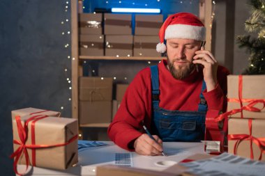 salesman in a Santa Claus hat takes an order by cell phone while sitting in an office with boxes of gifts and surprises for Christmas for office workers. Shopping and delivery. Small business concept