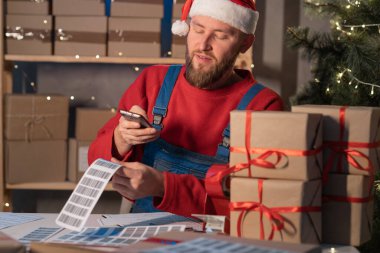 Young Santa Claus businessman sitting at his desk and scanning a delivery box with his mobile phone. Man selling products online on Christmas, scanning the barcode with his smart phone.