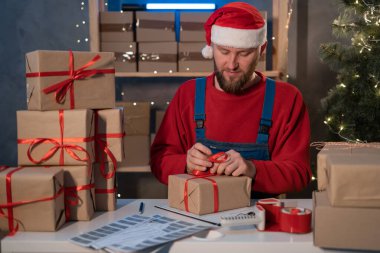 bearded man wearing a Santa Claus hat works in a warehouse of boxes with gifts and orders from an online store for Christmas. Small Business Owner Sells Happy New Year. Delivery holiday.