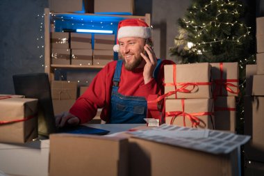 salesman in a Santa Claus hat takes an order by cell phone while sitting in an office with boxes of gifts and surprises for Christmas for office workers. Shopping and delivery. Small business concept