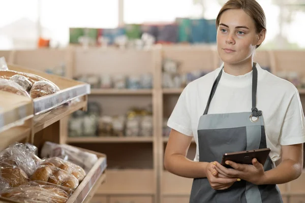 Attractive female staff using digital tablet in supermarket, woman wearing uniform with apron working in grocery store holding tablet pc