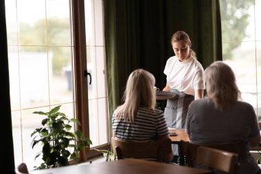 Young happy waitress serving coffee while talking to female guests in a cafe. copy space