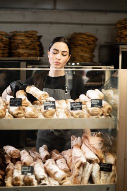 Beautiful young woman working in a bakery shop, bakery owner at work, showcase with pastry assortment with price tags, small business concept