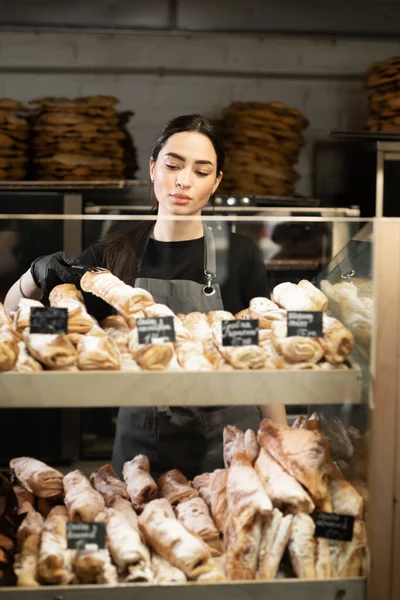 Beautiful young woman working in a bakery shop, bakery owner at work, showcase with pastry assortment with price tags, small business concept
