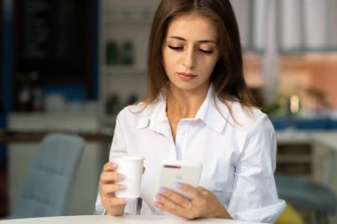 Charming young woman reading good news on mobile phone while relaxing in cafe, happy caucasian girl looking at photos on cell phone in free time in coffee house