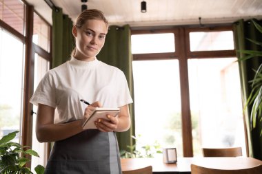 smiling waitress in black apron ready to take customer order, attractive young woman with notepad and pen looking at camera, standing in cozy coffee shop, good service concept