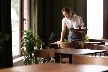 Waitress cleaning a table in a restaurant, taking away cups on a tray, hospitality service concept