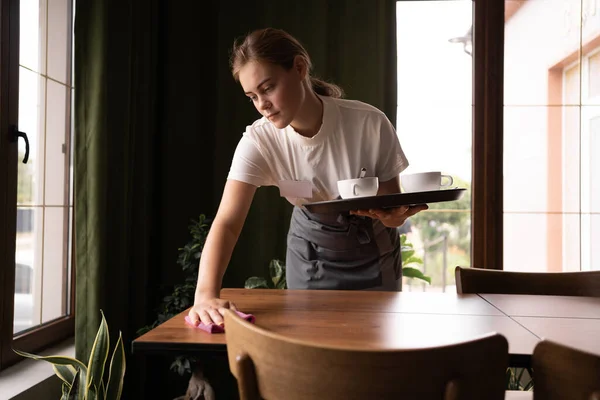 Waitress cleaning a table in a restaurant, taking away cups on a tray, hospitality service concept