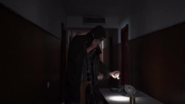 a man in clothes with a hood and a gas mask, walking along an old abandoned corridor with a flashlight, finds canned food in the dust. Dystopian concept, future, survivor. Post-apocalyptic world.