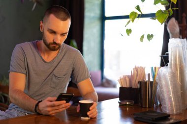 Successful young businessman is using a smartphone and drinking coffee while sitting at bar counter in a modern urban cafe. Technologies make life easier.