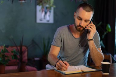Confident bar owner. handsome young male bartender leaning at the bar counter writing something in note pad and have call on mobile phone while sitting at the cafe