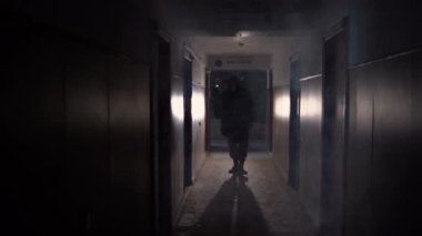 Man in respirator gas mask, hood and camo pants walk on the inside of abandoned building with flashlight. Cyberpunk postapocalypse fantasy horror scene or air pollution