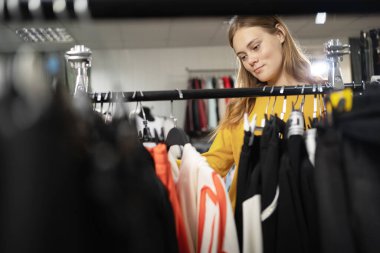 Genç bir kız spor giyim mağazasından kıyafet seçiyor. Eşofman altı, ceket ve askıda asılı bir sweatshirt. Kadın kıyafetleri, gençlik giysileri. Boşluğu kopyala