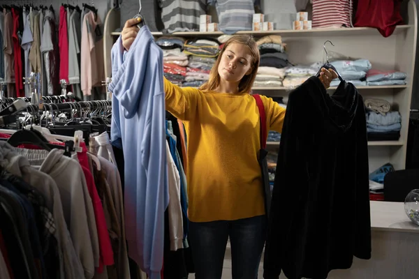 happy young woman choosing between two sweatshirts in mall or clothing store. sale, clothing, shopping, fashion and people concept