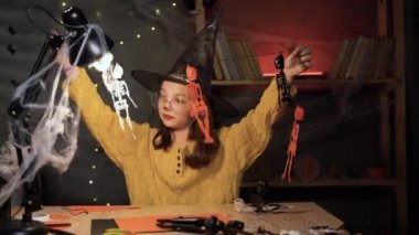 Little girl dressed as a witch preparing Halloween decor. Sitting at the table child makes a garland of skeletons. DIY Halloween concept