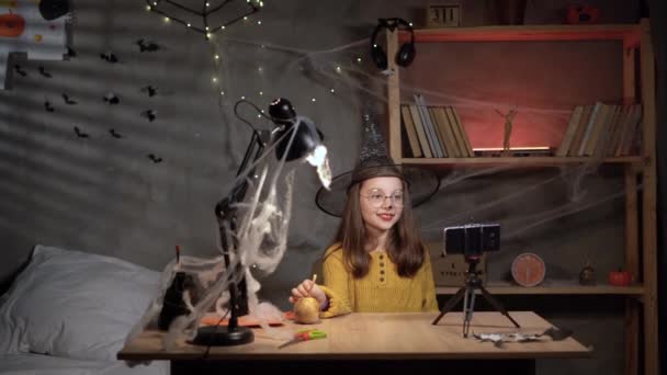 小さな女の子ブロガーでハロウィン魔女帽子レコーディングビデオのためにブログ上のスマートフォン リモートハロウィンの概念 — ストック動画