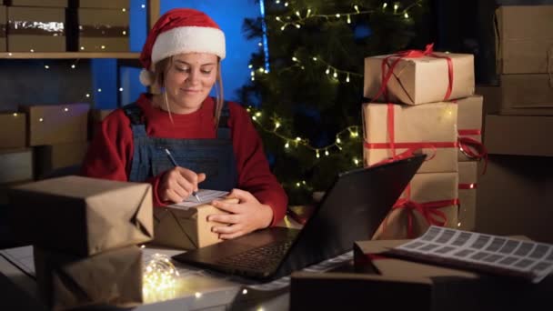 クリスマスの家のオフィスから働く女性の小さいビジネス所有者は夕方のラップトップを使用してオンラインで注文を取っている間箱の住所を書くことを前夜 ビッグクリスマスオンライン販売 — ストック動画