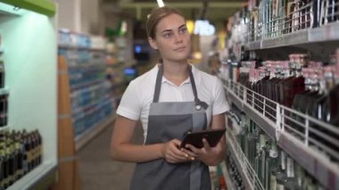 Sıradan gri önlüklü bir mağazada başarılı bir işçi süpermarkette alkolle rafların arasında yürüyor ve elinde bir tablet tutuyor. Genç bayan stajyer konsepti.