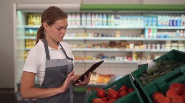 Young woman, employee with apron working on digital tablet and checking condition of product in the store, standing near vegetables shelves, Friendly smile at the camera.