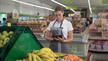 Young woman supermarket employee with apron working on digital tablet and checking condition of product in the grocery store. Fruit department of shop
