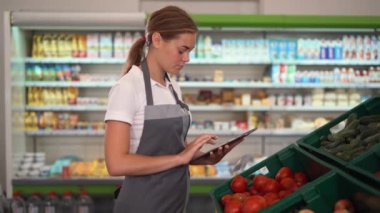Young woman, employee with apron working on digital tablet and checking condition of product in the store, standing near vegetables shelves, Friendly smile at the camera.