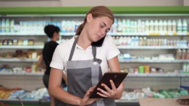 Close up of female seller in shop accepting order on mobile phone while standing near shelves. Young woman holding digital tablet in hand in grocery store.