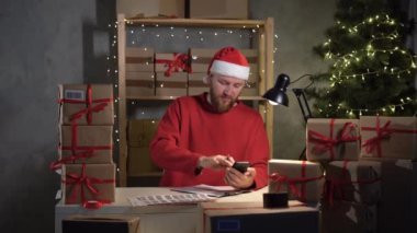 Christmas present delivery small business owner, man wearing santa claus hat working with boxes in home office taking online order using mobile phone, SME, e-commerce, telemarketing holiday concept