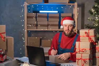small business startup owner Santa Claus working at home with laptop. Online Christmas sale. man sits at a table with boxes and writes the delivery address. Holiday SME concept