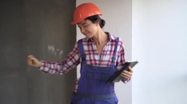 Şantiyede dans eden mutlu ve heyecanlı kadın inşaat işçisi üniforma ve güvenlik kaskı giyip elinde dijital tabletle eğleniyor.