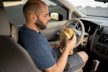 Adam araba sürerken hamburger yiyor. Trafikte yemek yerken arabasını süren aç bir sürücü.
