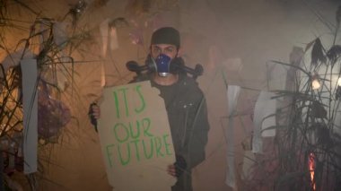 Gaz maskeli bir adam elinde bizim geleceğimizle ilgili kelimelerle poster tutuyor. Arka planda yanan plastik çöpler. Çevresel sorunlar kavramı: plastik çöpler, yasadışı çöplükler, atık yakma