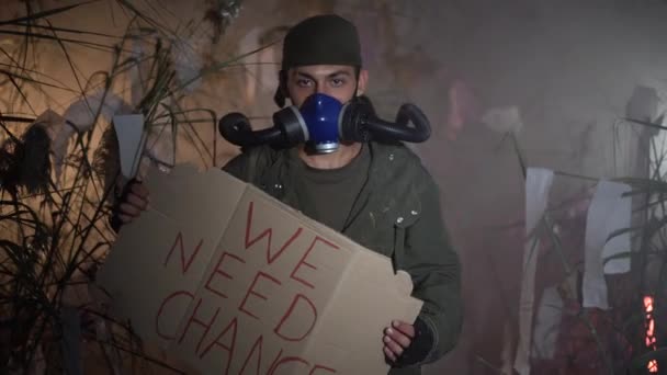 一个戴着防毒面具的男人举着写着 我们需要变革 字样的海报 在后面烧塑料垃圾 环境问题的概念 塑料垃圾 非法倾倒 废物焚化 — 图库视频影像