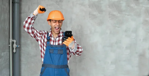 Photo of a happy positive young man builder in helmet posing isolated over gray wall background using mobile phone. Human positive emotion concept. banner