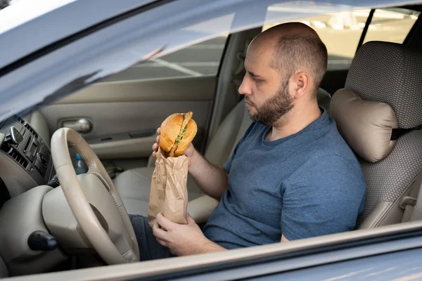 Man driving car while eating hamburger. Waiting and standing in traffic jam on road
